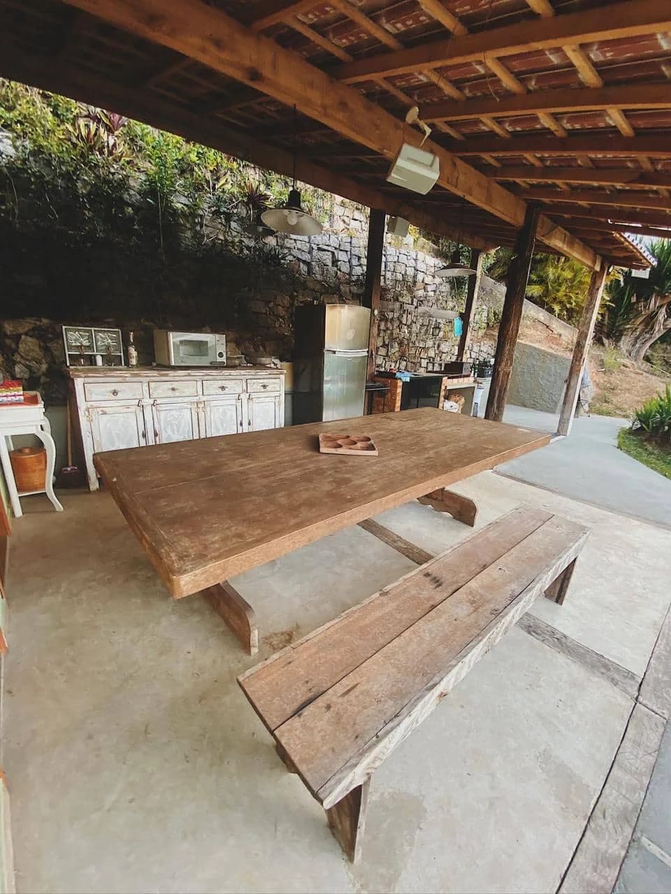 A churrasqueira coberta, com bancada, geladeira e uma mesa comprida de madeira maciça com bancos